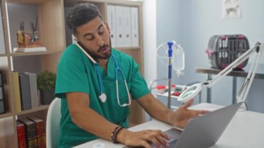 Yeşil önlüklü genç bir İspanyol erkek veteriner, iyi donanımlı bir klinikte çalışıyor, telefonda ve laptopta birden fazla iş yapıyor, hareketli ve verimli bir tıbbi ortam sergiliyor..