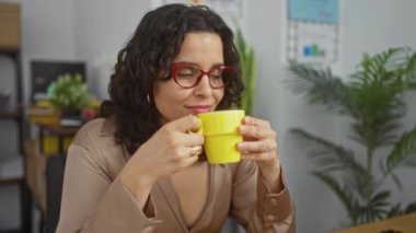 Ofis ortamındaki genç ve güzel bir İspanyol kadın sıcak bir içecekten zevk alıyor, rahat ve memnun görünüyor, çevresi kapalı bitkiler ve işyeri dekoruyla çevrili..