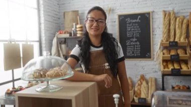 Bir pastanede çalışan, gözlüklü, fırında pişirilmiş yiyeceklerle poz veren ve modern kapalı bir ortamda gülümseyen bir kadın.