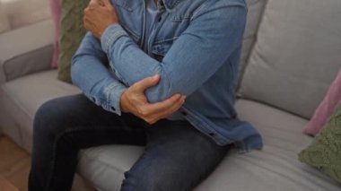 Acı dolu dirseğini bir salonda tutan, kot ceket giyen, rahatsızlık veren, ev dekoruyla çevrili bir adam, evde kol yaralanmasından muzdarip yetişkin bir erkek olduğunu gösteriyor.