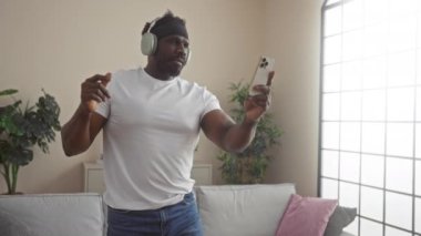 Genç, yakışıklı, Afro-Amerikan bir adam evde, oturma odasında, kulaklık takmış ve elinde akıllı bir telefonla video görüşmesi yapıyor..