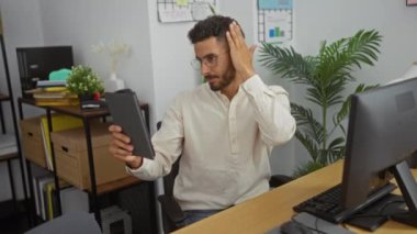 Ofisteki genç bir İspanyol, ofis mobilyaları, bitkiler ve belgelerle çevrili bir tabletle selfie çekerken saçını düzeltiyor..