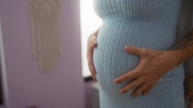 Sıcak bir evde elleri karnında kucaklaşan hamile çift.