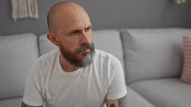 Modern bir oturma odasında oturan sakallı ve dövmeli yakışıklı adam gri kanepeli ve pembe yastıklı.