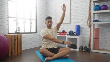 Genç İspanyol bir adam arka planda çeşitli fitness ekipmanlarıyla iyi donanımlı bir spor salonunda yoga minderinde esneme egzersizleri yapıyor..