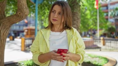 Şehir parkında elinde kırmızı bir telefonla güneşli bir günde açık havada dikilen beyaz ve sarı giyinmiş güzel sarışın beyaz bir kadın..