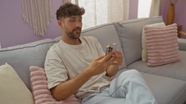 Evdeki gri bir kanepede rahat bir oturma odasında akıllı telefon kullanan genç bir adam.