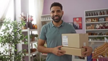 Paketleri olan genç bir adam, arka planda bitki ve rafları olan bir ev dekoratörü dükkanında duruyor.