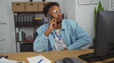 Genç bir Afrikalı Amerikalı adam, elinde telefon olan modern bir ofiste masasında oturuyor. Etrafı ofis malzemeleri ve belgelerle çevriliyken düşünceli görünüyor..