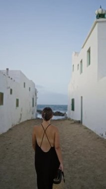 Beyaz binalar arasında kumlu bir sokakta yürüyen genç bir kadın. Aile, lanzarot, kanarya adaları, siyah bir elbise giymiş ve elinde hasır bir şapka tutuyor.