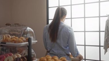 Pastanede çalışan genç bir kadın hamur işleri ve çöreklerle dolu bir vitrinin yanında duruyor, önlük giyiyor ve büyük bir pencerenin önünde hafif ışıkla gülümsüyor.