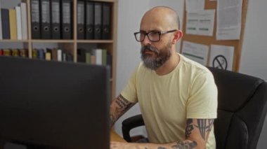 Ofis odasında bilgisayar üzerinde çalışan, gözlüklü ve dövmeli yetişkin erkeği vurgulayan, sakallı İspanyol bir adam, dosya ve kitapların arasındaki ekrana odaklandı..