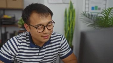 Genç Çinli bir adam, modern bir işyerinde bitki ve raflarla çevrili kapalı bir ofis ortamında çalışırken gülümsüyor..