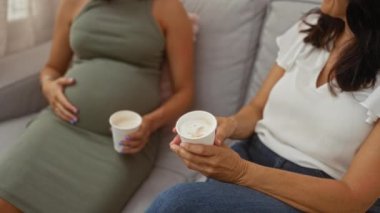 Hamile kadın ve anne sıcak bir oturma odasında kahvelerini yudumluyor, gülümsüyor ve sohbet ediyorlar.