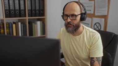 Sakallı ve dövmeli İspanyol bir adam kulaklık takıyor ve gözlük takıyor. Arka planda kitap ve klasörlerle dolu raflarla modern bir ofiste çalışıyor..