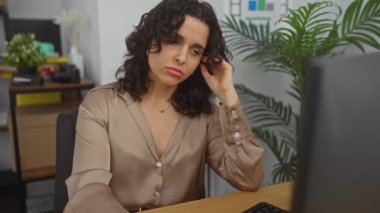 Ofis ortamında masasında çalışırken esneyen ve yorgun görünen İspanyol kadın. Etrafı kapalı bitkiler ve ofis malzemeleriyle çevrili.