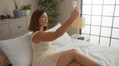 Yatak odasında dinlenen bir kadın elinde akıllı telefonuyla selfie çekerken odada beyaz çarşaflar ve çeşitli bitkilerle bir yatakta oturuyor.