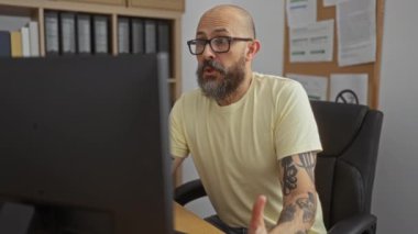 Ofisteki bilgisayarda çalışan, kolları dövmeli arka planda kitap rafı ve duvarında ilan tahtası olan bir adam.