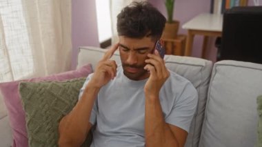 Genç bir adam endişeli bir ifadeyle oturma odasındaki bir koltukta oturmuş telefonla konuşuyor ve şakağına işaret ediyor. Stres ya da hayal kırıklığını işaret ediyor.