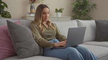 Sarı saçlı genç bir kadın rahat bir koltukta oturuyor. Kapalı bir şehir ortamında dizüstü bilgisayar kullanıyor. Etrafında bitkiler ve yumuşak yastıklar var..