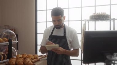 Fırında çalışan genç adam tezgahın arkasında notlar alıyor. Pasta dolu, önlük takıyor, kapalı bir dükkânda büyük pencerelerden gelen doğal ışıkla.