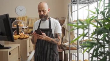 Fırında telefon kullanan ve arka planda ekmek ve hamur işleri olan bir adam.
