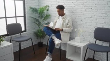 Genç Afro-Amerikalı bir adam kapalı bir bekleme odasında oturmuş, bitkiler ve minimalist mobilyalar da dahil olmak üzere modern dekorlarla çevrili bir tablet okuyor..
