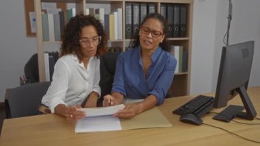 Ofiste çalışan iki kadın, belgeleri inceliyor ve bilgisayarın önünde tartışıyorlar. İşbirlikçi ve profesyonel çalışma ortamını vurguluyorlar..
