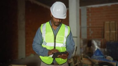 Güvenlik yeleği giyen genç adam bitmemiş bir inşaat alanında tablet üzerinde çalışıyor.