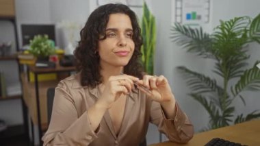 Genç, çekici esmer bir kadın ofis ortamında, kapalı bir yerde, bitkiler ve dekorla çevrili, düşünceli bir poz veriyor..