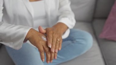 Orta yaşlı bir kadın sıcak bir oturma odasında kanepede otururken ellerine krem sürüyor.