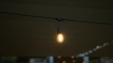 Işıldayan, açık hava ampulü loş ışıklandırılmış bir ortamda bir ipten sarkıyor ve arka planda bulanık ışıklar ve ılık bir ortam yaratıyor..