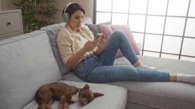 Genç bir kadın oturma odasında bir kanepede dinleniyor, kulaklıkla müzik dinliyor, akıllı bir telefon tutuyor, yanında bir köpek uyuyor..