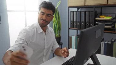 Arka planda yeşil bitki ve bilgisayar olan genç bir adam büro masasında selfie çekiyor.