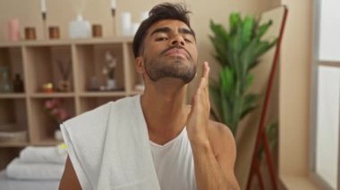Genç, yakışıklı, spa merkezindeki İspanyol bir adam, sakalına dokunuyor, omuzlarına havlu sarılı, kapalı bir yerde dikiliyor.
