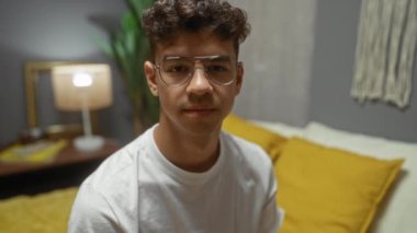 Gözlüklü genç İspanyol bir adam, rahat bir yatak odasında, rahat ve sakin bir ifade sergiliyor..