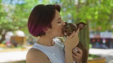 Kısa saçlı ve beyaz tişörtlü genç bir kadın güneşli bir günde açık hava parkında köpeğini sevgiyle tutuyor ve öpüyor..