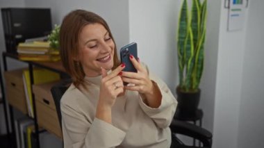 Ofiste akıllı telefon kullanan, gülümseyen ve duygularını ifade eden çekici genç sarışın kadın arka planda bitki ve ofis malzemeleriyle modern kapalı bir iş ortamında oturuyor..