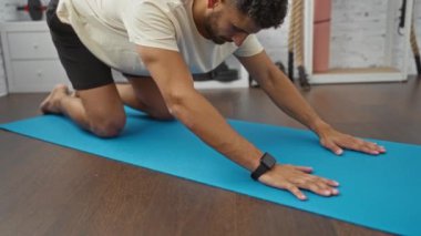 Latin kökenli genç bir adam spor salonundaki mavi yoga minderinin üzerinde esneme egzersizi yapıyor..