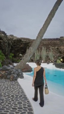 Lanzarote 'daki Jameos del agua' da volkanik kayalarla çevrili, güneş gözlüğü takan ve arkasında tropik bitkiler olan bir el çantası taşıyan bir kadın.