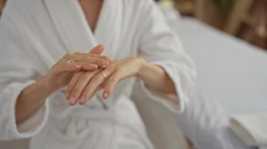 Kaplıca odasında el kremi süren bir kadın. Alyansla narin parmaklarını sergiliyor. Kapalı alandaki güzellik ya da sağlık seansının göstergesi olarak beyaz bir bornoz giyiyor..