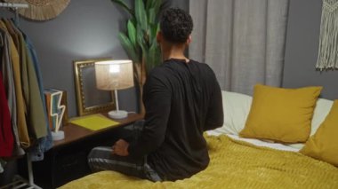 Genç adam rahat bir yatak odasında uzanır sarı yastıklar ve battaniyeyle, elbise askısı ve dekorla çevrili..