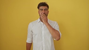 Beyaz tişörtlü, esneyen, sarı arka plana yaslanan, yorgunluk ve yorgunluk gösteren İspanyol genç bir adam.