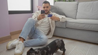 Akıllı telefonu ve kredi kartıyla oturma odasında köpeğiyle oturan genç bir İspanyol.
