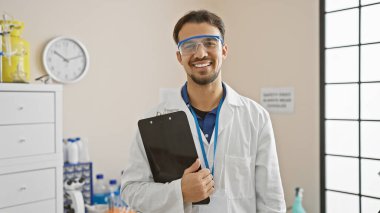 Laboratuvar önlüğü ve güvenlik gözlüğü takan sakallı, labaratuvarda pano tutan genç İspanyol bir adam.