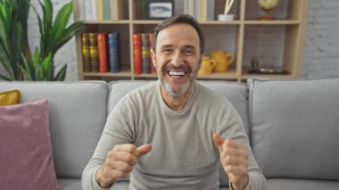 Sakallı, neşeli, orta yaşlı bir adam. Oturma odasında gülümseyip el sallıyor..