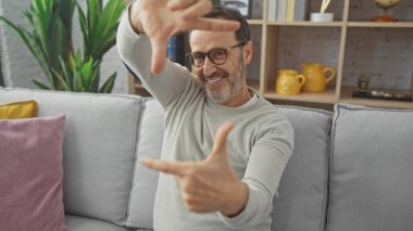 Gülümseyen orta yaşlı, sakallı, elleri rahat oturma odasında çerçeveli bir adam..