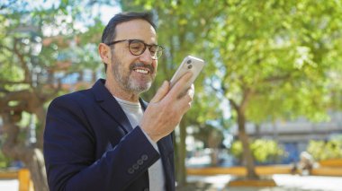 Sakallı, yakışıklı, olgun bir adam güneşli bir parkta akıllı telefon kullanıyor. Açık havada modern teknolojiyi temsil ediyor..