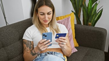 Young woman comparing credit card to phone app, sitting indoors on a sofa with houseplants in the background. clipart