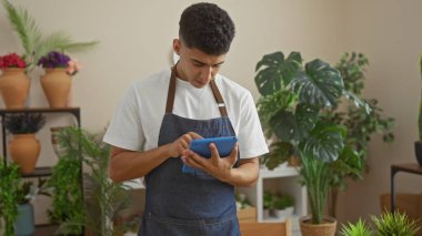 Önlük giyen genç bir adam, çeşitli bitkilerle çevrili kapalı bir çiçekçide tablet kullanıyor..